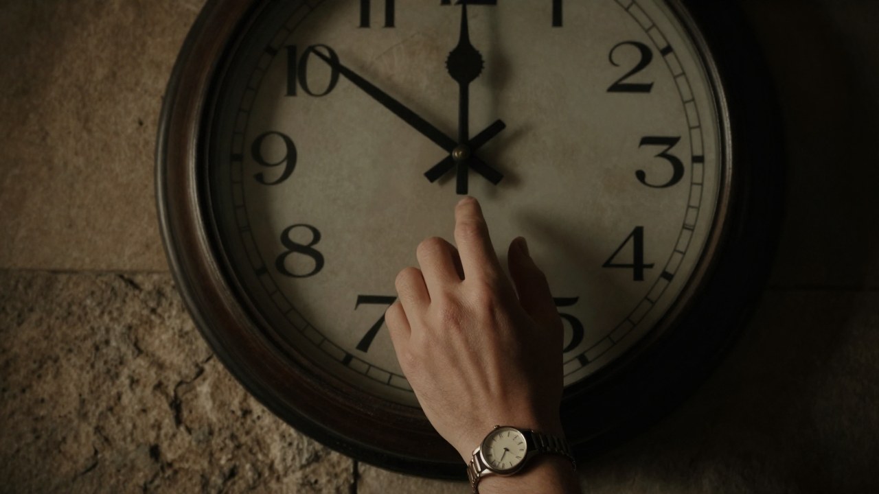 Reloj antiguo de pared con manecillas detenidas junto a una mano que lleva un reloj de pulsera.